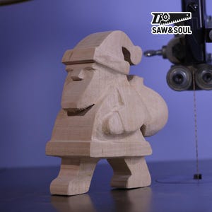 May include: A wooden Santa Claus figure, with a sack over his shoulder, is positioned in front of a band saw. The figure is made of light-coloured wood and has a simple, blocky design. The logo "SAW & SOUL" is in the upper right corner.