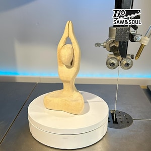 Könnte beinhalten: Eine hellfarbene Holzskulptur einer Person in einer Yoga-Pose, mit erhobenen Armen. Die Skulptur steht auf einer weißen Drehplattform. Das Bild zeigt auch eine Bandsäge und den Text "SAW&SOUL".