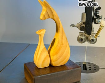 May include: Two wooden sculptures, one tall and curved, the other smaller, sit on a dark wooden base. The sculptures are light-colored with visible wood grain. The text "SAW&SOUL" is visible in the upper right corner.
