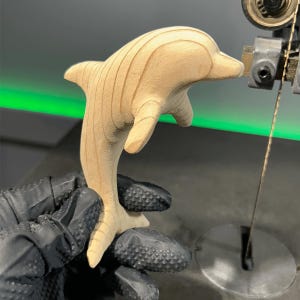May include: A wooden dolphin sculpture is being cut out on a band saw. The dolphin is light brown and has a smooth finish. The saw blade is visible in the background.