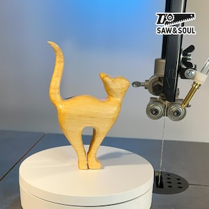 Puede incluir: Una figura de gato de madera con un acabado liso y de color claro. El gato está de pie con la cola curvada hacia arriba. La figura está sobre una plataforma giratoria blanca, con una sierra de cinta en el fondo. El logo "SAW&SOUL" es visible.
