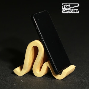 Könnte beinhalten: Ein hellfarbener Handyhalter aus Holz mit wellenförmigem Design, der ein schwarzes Smartphone hält. Der Ständer ist aus Naturholz gefertigt und trägt den Text "Saw & Soul" in der oberen rechten Ecke.