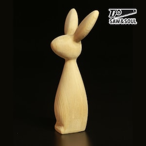 Op de afbeelding: Een houten konijnbeeldje met lange oren en een gladde, natuurlijke afwerking. De sculptuur is gesneden uit lichtgekleurd hout en staat rechtop op een platte basis. Het logo "SAW&SOUL" staat in de rechterbovenhoek.