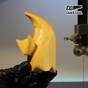 Könnte beinhalten: Eine geschnitzte Holzgeisterfigur mit geschwungenem Oberteil und einer kleinen, flügelförmigen Form an der Seite. Das Holz hat eine helle, natürliche Farbe und eine glatte Oberfläche. Die Figur wird in einer schwarzen behandschuhten Hand gehalten.
