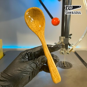 May include: A handcrafted wooden spoon with a long handle and a rounded bowl. The spoon is made of light-colored wood with visible grain patterns. The spoon is held by a gloved hand. The logo "SAW & SOUL" is visible in the background.