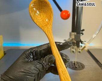 May include: A handcrafted wooden spoon with a long handle and a rounded bowl. The spoon is made of light-colored wood with visible grain patterns. The spoon is held by a gloved hand. The logo "SAW & SOUL" is visible in the background.