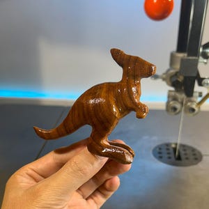 Puede incluir: Figurita de canguro de madera hecha a mano con un acabado brillante. El canguro es marrón con vetas de madera visibles y está en posición de pie. La figura se sostiene en la mano de una persona.