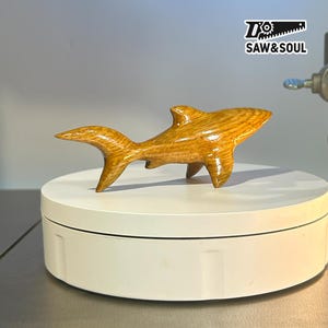 May include: A handcrafted wooden shark figurine with a glossy finish. The shark is carved from light brown wood, showcasing the natural grain. The figurine is displayed on a white rotating platform. The logo "SAW&SOUL" is visible in the top right corner.