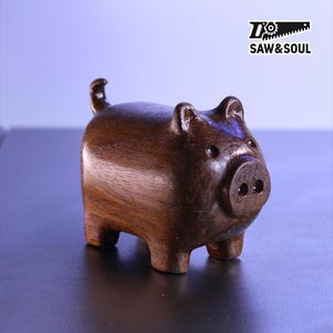 May include: A handcrafted wooden pig figurine with a smooth, polished finish. The pig is a rich brown color and has a small curly tail. The figurine has a detailed snout and small ears. The logo "SAW&SOUL" is in the upper right corner.