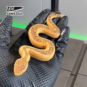 May include: A hand wearing a black glove holds a wooden snake sculpture. The snake is carved with detailed scales and has a light brown color.