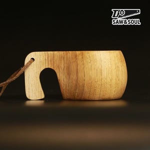 Op de afbeelding: Een lichtbruine houten kuksa-beker met een gladde afwerking en een gebogen handvat met een bruin leren koord. Rechtsboven staat een wit logo met een zaagpictogram en de tekst SAW&SOUL. De beker heeft een duidelijk zichtbare houtnerf.
