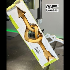Puede incluir: Una flecha de madera con un nudo en forma de corazón se está cortando en una sierra de calar. La madera es de color marrón claro y la sierra es de color plateado. La sierra está etiquetada como "Saw & Soul".