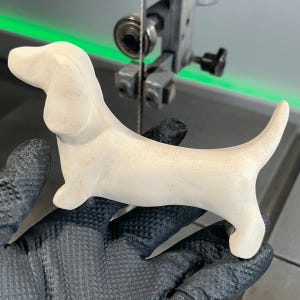 May include: A light-coloured, wooden dachshund dog figurine. The dog is carved with smooth lines and has a long body, short legs, and a curved tail. The figurine is held by a gloved hand, with a bandsaw in the background.