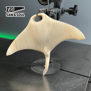 Peut inclure: Une sculpture de raie manta en bois en cours de découpe sur une scie à chantourner. La sculpture est en bois clair et est en cours de découpe. La scie est en arrière-plan et est partiellement visible.