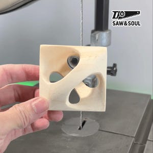 May include: A light-coloured wooden cube with curved cutouts is held near a band saw. The cube has a unique design with flowing, organic shapes carved into its sides. The logo "SAW & SOUL" is visible in the upper right corner.