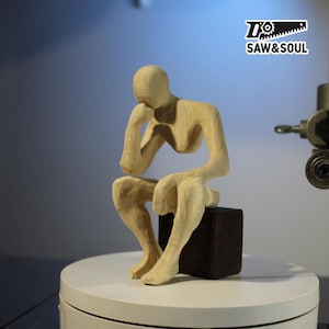 Può includere: Scultura in legno chiaro di una figura seduta, con la testa appoggiata su una mano, che suggerisce contemplazione. La figura è posizionata su una base quadrata marrone scuro. La scultura è esposta su una superficie bianca.