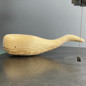 Puede incluir: Una escultura de madera de una ballena que se está cortando en una sierra de calar. La ballena está hecha de madera clara y tiene una forma lisa y redondeada.