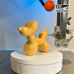 May include: A wooden balloon dog sculpture with a light brown finish. The dog is posed on a white rotating platform. The logo "SAW & SOUL" is visible in the background.