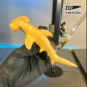 May include: A wooden hammerhead shark sculpture, crafted from light-coloured wood. The shark's natural grain is visible. The sculpture is held by a gloved hand, with the "SAW&SOUL" logo in the background.