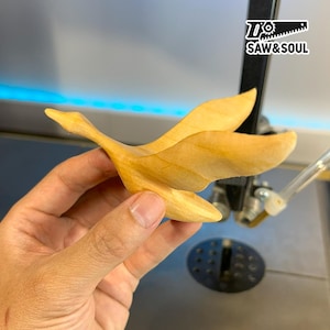 Può includere: Una scultura in legno di un'oca in volo, realizzata in legno di colore chiaro. L'uccello è raffigurato a mezz'aria con le ali spiegate e un lungo collo. Il logo "SAW&SOUL" è visibile sullo sfondo.
