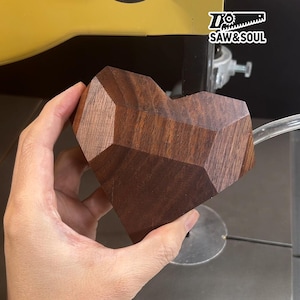 Könnte beinhalten: Ein facettiertes Holzherz, gehalten in einer Hand, mit einer tiefbraunen Farbe. Das geometrische Design des Herzens ist erkennbar, mit dem Logo "SAW & SOUL" oben rechts. Die Holzmaserung ist sichtbar, was die Handwerkskunst hervorhebt.