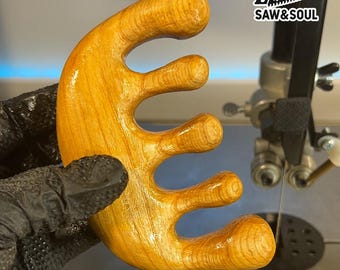 May include: A handcrafted wooden comb with a unique, ergonomic design. The comb is made of light-colored wood with a visible grain and a smooth, polished finish. The comb has a curved handle and several rounded teeth. The logo "SAW&SOUL" is visible in the background.