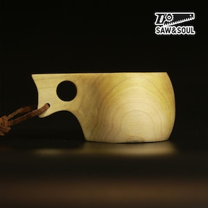 May include: A light-colored wooden kuksa cup with a smooth, rounded bowl and a handle featuring a circular finger hole. A brown leather cord is attached to the handle. The top right corner displays the white text logo SAW&SOUL with a saw icon.