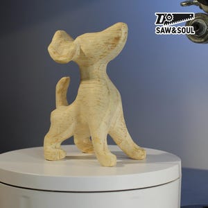 Może przedstawiać: Drewniana rzeźba psa o gładkim, jasnym wykończeniu. Pies jest wyrzeźbiony w stylizowanej pozie, z głową uniesioną do góry i lekko uniesionym ogonem. Rzeźba stoi na białej powierzchni, z logo "Saw & Soul" w prawym górnym rogu.