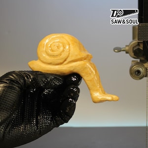 Könnte beinhalten: Eine handgeschnitzte Holzschnecken-Skulptur mit einem spiralförmigen Gehäuse und einem einzelnen Bein. Die Schnecke ist hellbraun und wird von einer behandschuhten Hand gehalten. Das Logo "SAW&SOUL" befindet sich in der oberen rechten Ecke.