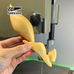May include: A wooden carving of a cartoon roadrunner in flight. The carving is light brown and has a smooth finish. The carving is being held in a hand in front of a band saw.