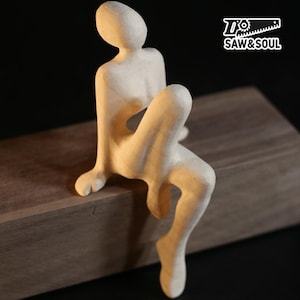 May include: A light-colored, carved wooden sculpture of a seated figure. The figure is positioned on a dark wooden block. The sculpture has a smooth, unpainted finish. The logo "SAW&SOUL" is visible in the upper right corner.