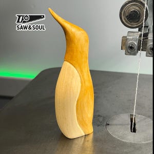 May include: A wooden penguin figurine with a light brown body and a white belly. The figurine is standing on a gray surface. The text "SAW & SOUL" is visible in the background.
