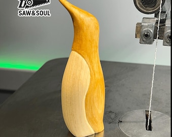 May include: A wooden penguin figurine with a light brown body and a white belly. The figurine is standing on a gray surface. The text "SAW & SOUL" is visible in the background.