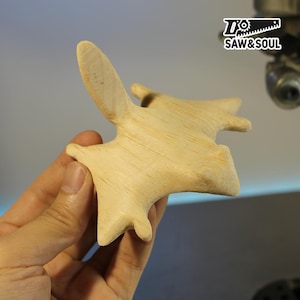 May include: A light-coloured wooden carving of a stylized bat or similar creature, held in a hand. The carving has a smooth finish and a wing-like extension. The logo "SAW&SOUL" is visible in the upper right corner.