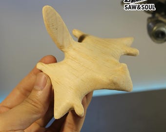 Könnte beinhalten: Eine hellfarbene Holzschnitzerei einer stilisierten Fledermaus oder eines ähnlichen Wesens, die in einer Hand gehalten wird. Die Schnitzerei hat eine glatte Oberfläche und eine flügelartige Verlängerung. Das Logo "SAW&SOUL" ist oben rechts sichtbar.