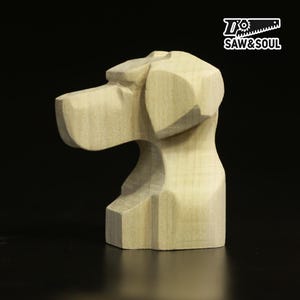 May include: A carved wooden dog head sculpture. The sculpture is light-coloured, with visible wood grain and geometric facets. The dog's head is in profile, with a prominent snout and ears. The logo "SAW&SOUL" is in the upper right corner.