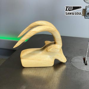 May include: A handcrafted wooden sculpture of an antelope head with curved horns. The sculpture is made of light-colored wood and is positioned on a metal surface. The logo "SAW & SOUL" is visible in the upper right corner.