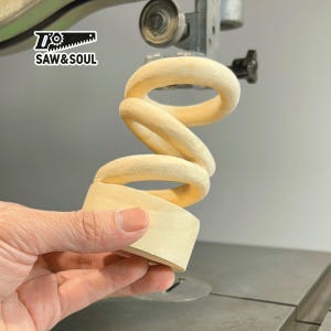 Könnte beinhalten: Eine Holzskulptur in Form einer Spirale wird auf einer Bandsäge geschnitten. Die Skulptur ist aus hellem Holz gefertigt und hat eine glatte Oberfläche. Die Säge ist grün und schwarz und trägt den Text "Saw & Soul".