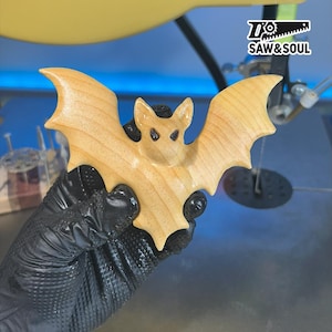 May include: A wooden bat figurine with a light brown finish. The bat has detailed wings and a carved face with dark eyes. The bat is held by a gloved hand. The logo "SAW&SOUL" is visible in the background.