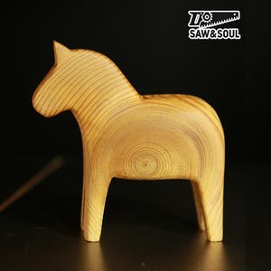 May include: A wooden horse figurine with a light brown color and visible wood grain. The horse has a simple, stylized design with a prominent head and a curved body. The logo "Saw & Soul" is in the top right corner.