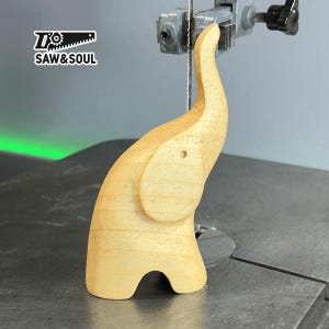 Puede incluir: Una figurita de elefante de madera que se está cortando en una sierra de cinta. El elefante es de color marrón claro y tiene un diseño sencillo. La sierra es una sierra de cinta negra y plateada con un fondo verde.