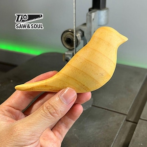 May include: A light wood bird carving, with a smooth, rounded shape and visible wood grain. The bird is held in a hand, with a green background behind it.