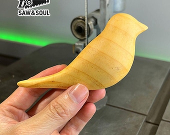 May include: A light wood bird carving, with a smooth, rounded shape and visible wood grain. The bird is held in a hand, with a green background behind it.