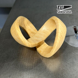 May include: A light-colored wooden sculpture in an infinity loop design. The wood grain is visible, and the piece is set against a metallic surface. The logo "SAW&SOUL" is in the upper right corner.