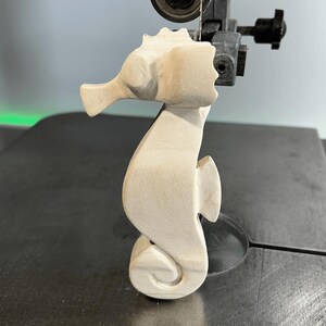 May include: A wooden seahorse sculpture being cut out on a band saw. The seahorse is unpainted and has a detailed design.