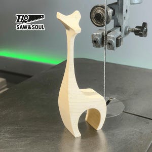 May include: A wooden giraffe sculpture being cut on a band saw. The giraffe is a simple design with a long neck and a rounded body. The saw blade is visible in the center of the image.