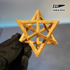 Peut inclure: Une sculpture géométrique en bois en forme d'étoile avec un design ouvert complexe. L'étoile est de couleur marron clair et est tenue par une main gantée. Le logo "SAW&SOUL" est dans le coin supérieur droit.