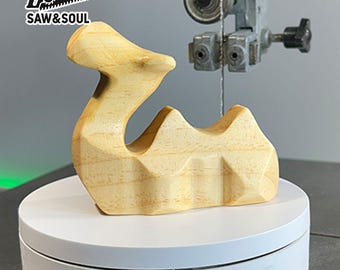May include: A light-colored wooden sculpture with a curved, abstract design. The sculpture has a smooth finish and geometric facets. The logo "SAW & SOUL" is in the upper left corner. The sculpture is on a white rotating platform.
