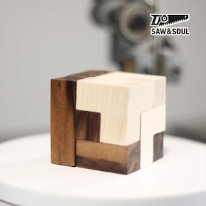 Könnte beinhalten: Ein Holzpuzzelwürfel mit ineinandergreifenden Teilen in hell- und dunkelbraun. Der Würfel steht auf einer weißen Oberfläche, im Hintergrund ist das Logo "SAW & SOUL" zu sehen.