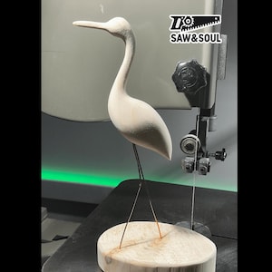 May include: A white wooden bird sculpture of a heron with black wire legs standing on a wooden base. The sculpture is being cut on a scroll saw with the text "Saw & Soul" on the machine.